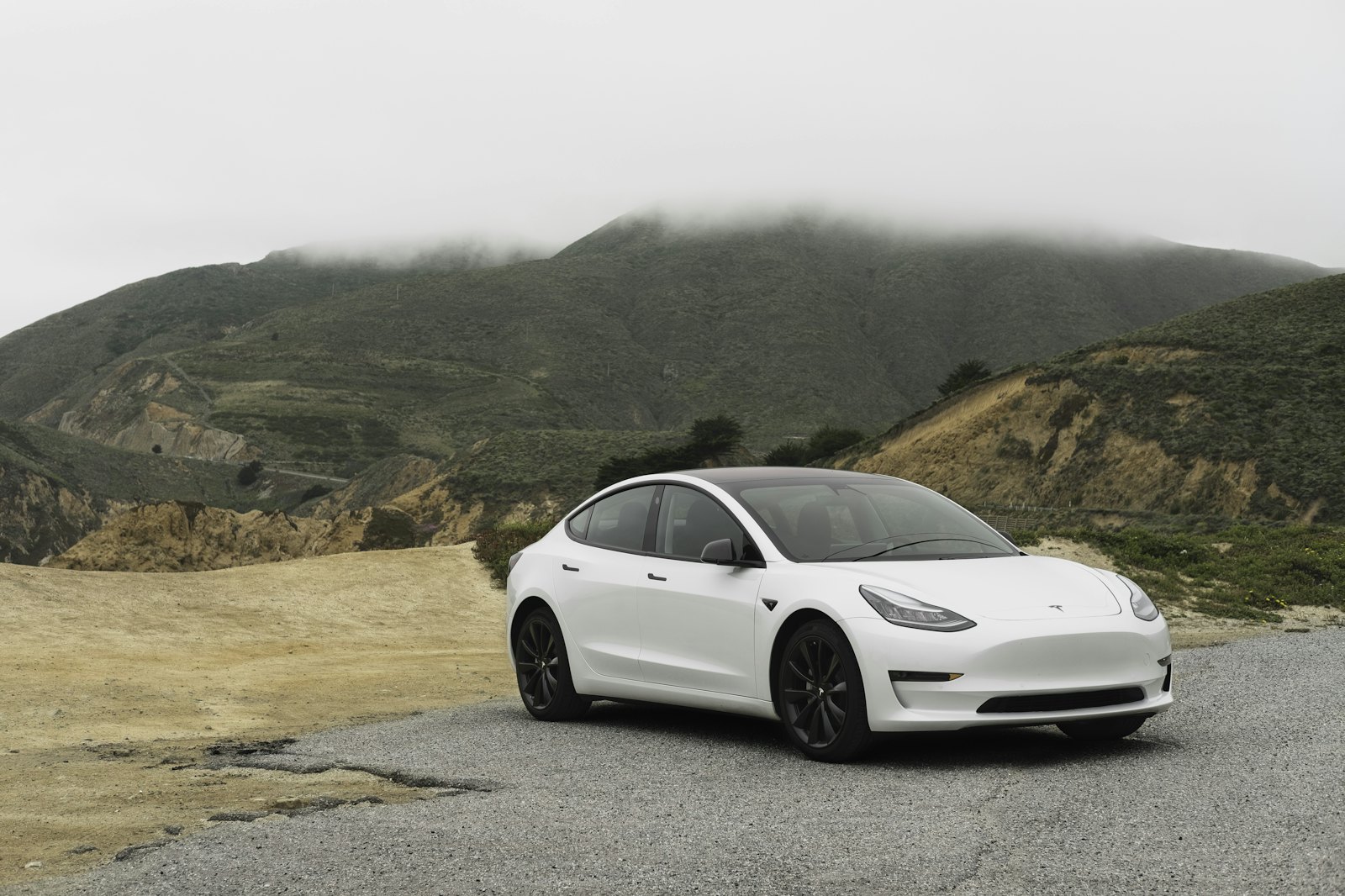 A white sedan parked on a gravel turnout below a foggy hill